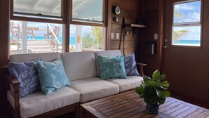 A wooden interior with a white sofa, two windows with a view of the beach, and a wooden table with a plant.