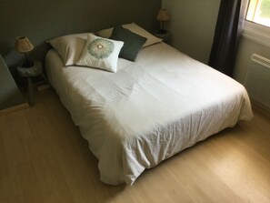 Una cama bien hecha con edredón blanco y un cojín decorativo, dos lámparas de noche y una ventana con cortinas.