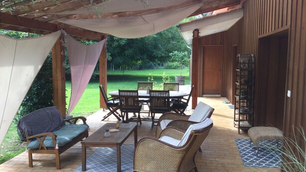 Eine Terrasse im Freien mit Holztisch und -stühlen, einer Korbschaukel und einem überdachten Bereich mit Blick auf einen grünen Garten.