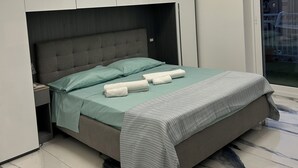 Un dormitorio moderno con cama, cabecero gris y vista a edificios a través de un ventanal grande.