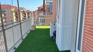 Un balcón con césped artificial verde, barandillas blancas y vistas a una zona residencial con edificios y cielo despejado.