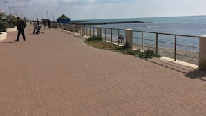 Un paseo pavimentado a lo largo de la costa, con barandilla, un edificio azul y gente caminando.