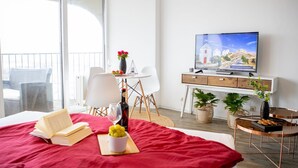 A hotel room with a bed, a desk with a TV, a balcony with a chair, and a dining area with a table and chairs.