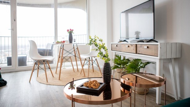 A modern living room with a glass coffee table, a flat-screen TV mounted on the wall, and a balcony with a dining area.