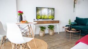 A modern living room with a white dining table, a white chair, a green sofa, a TV on a wooden stand, and two potted plants.