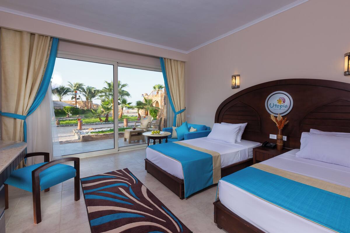 A hotel room with two beds, a desk, a chair, a small table, and a view of the outdoors.
