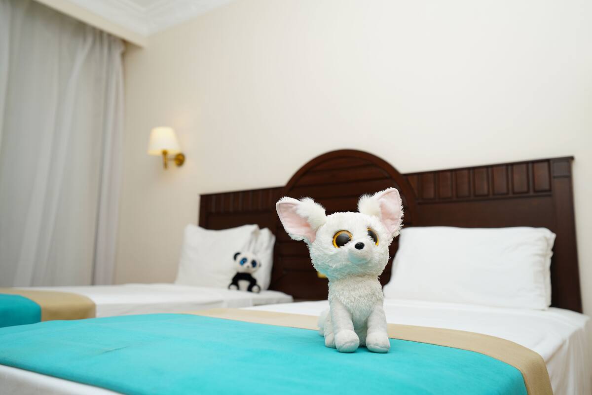 A plush toy on a bed with a wooden headboard, a lamp, and a panda plush toy in the background.