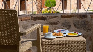 Una silla de mimbre y una mesa con alimentos para el desayuno, una palmera y vistas al mar y edificios de piedra.