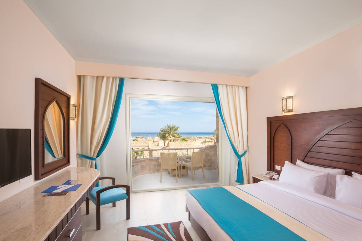 A hotel room with a bed, a desk, a chair, a TV, and a balcony with a view of the beach.