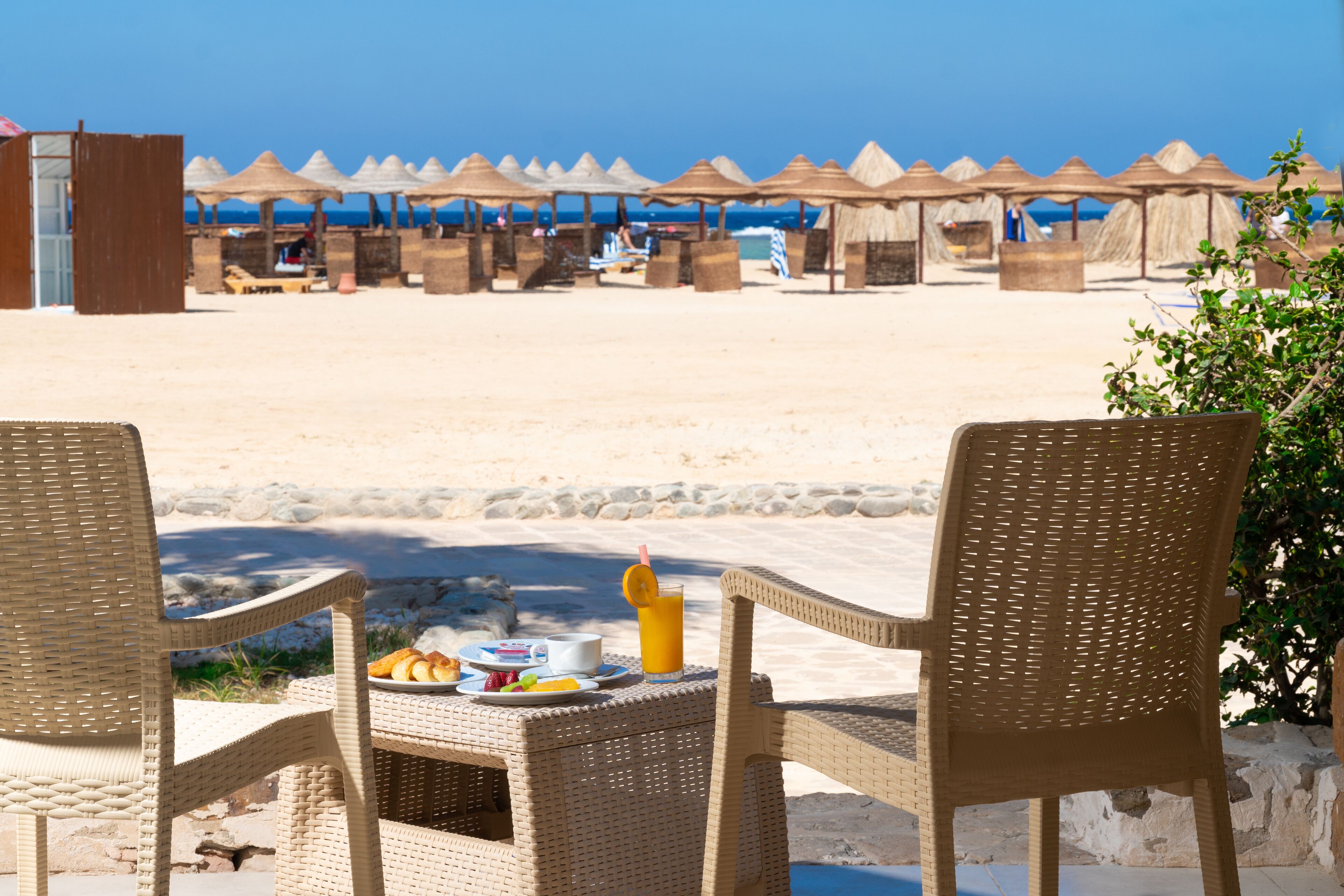 En matplats vid stranden med flätade stolar och ett bord dukat med frukostmat.