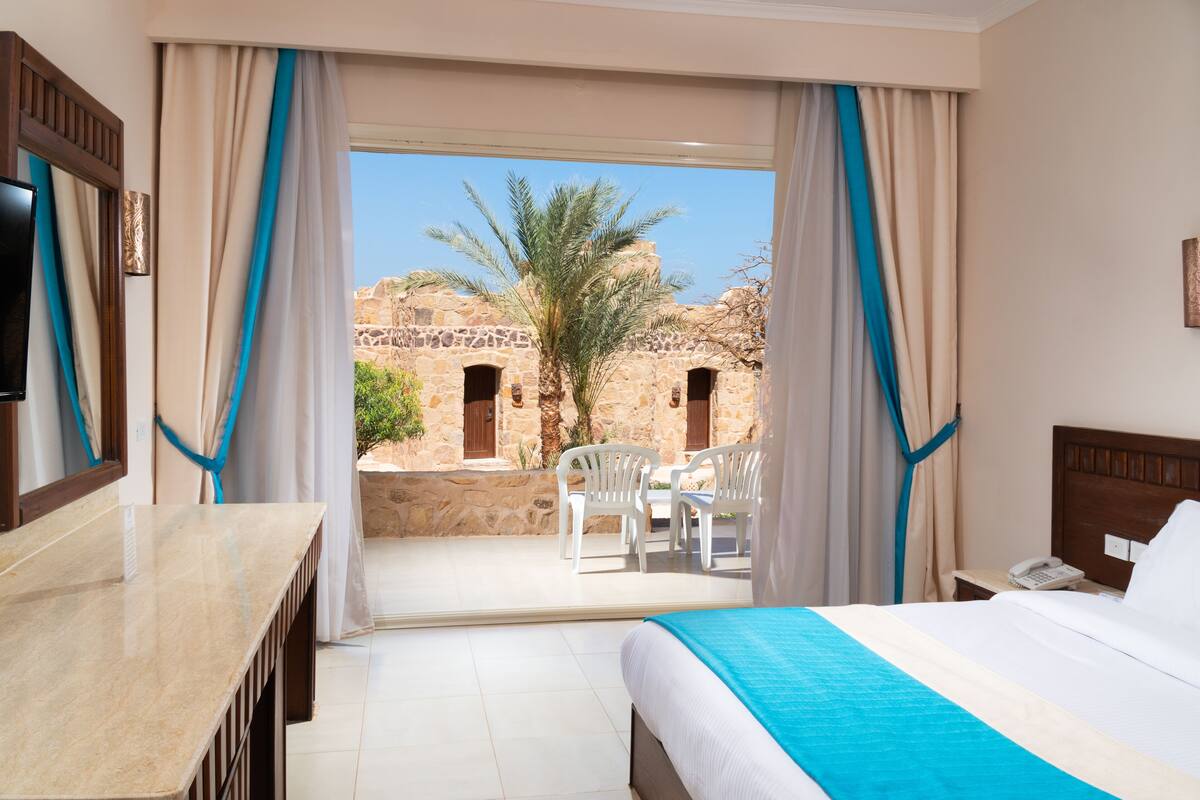 A hotel room with a bed, a desk, and a balcony with a view of a stone building and palm tree.