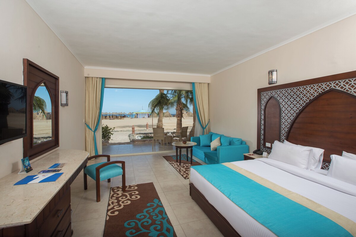 A hotel room with a bed, a desk, a chair, a small table, and a view of the beach.