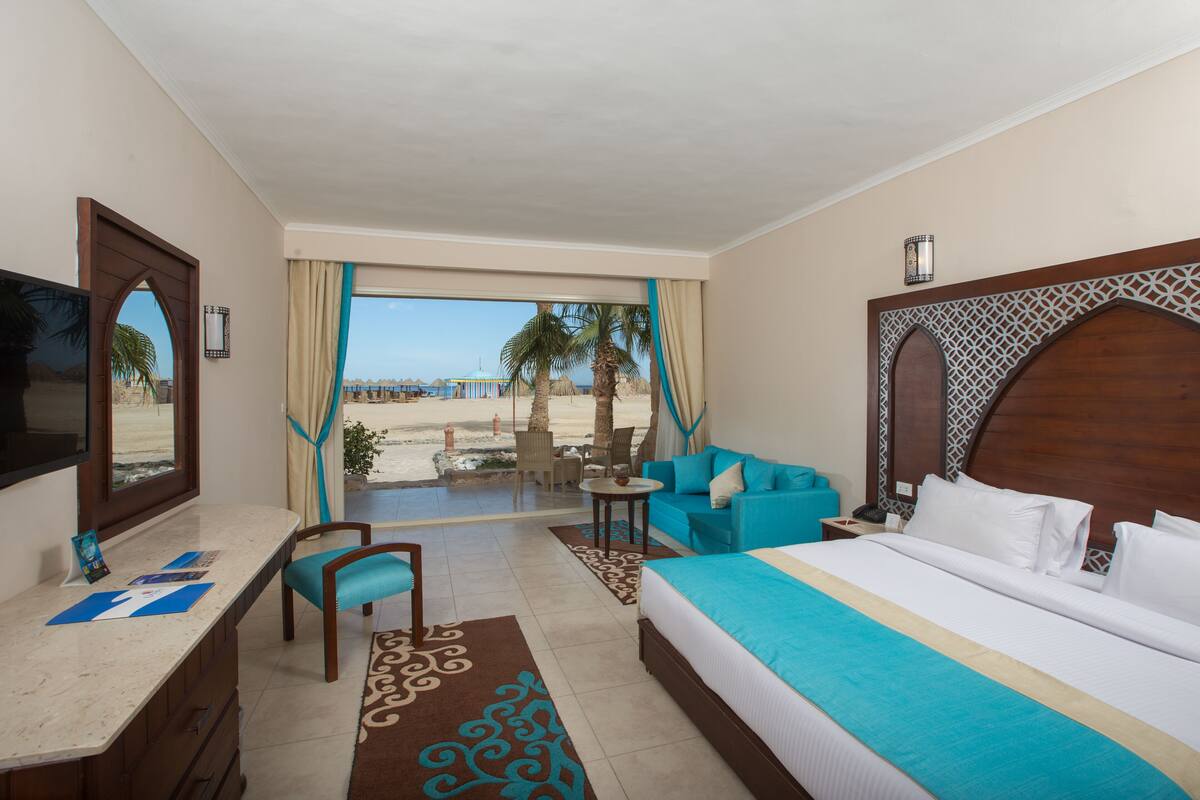 A hotel room with a bed, a desk, a chair, a small table, and a view of the beach.