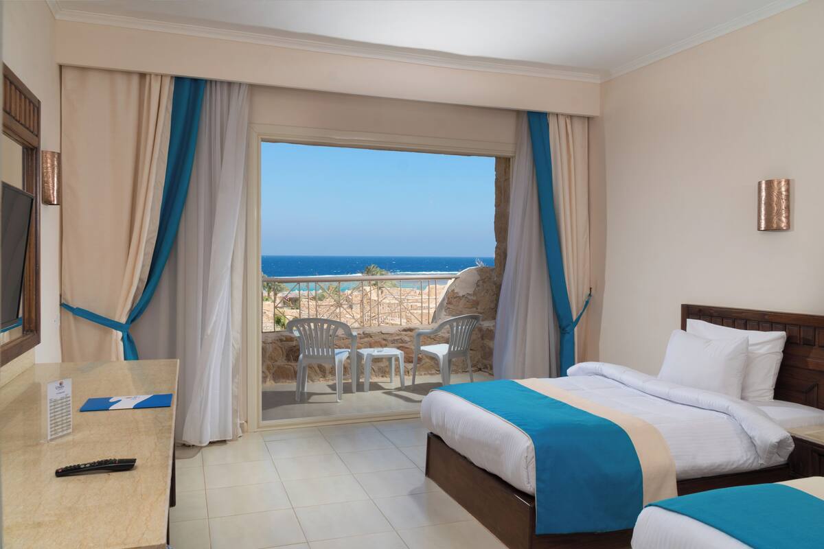 A hotel room with a bed, a desk, a TV, and a balcony with a view of the sea.