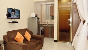 A living room with a brown sofa, a television, a kitchen area with a refrigerator, and a door leading to another room. - Studio Furnished Apartments Entebbe (Entebbe)