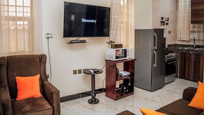 A living room with a flat-screen TV, a sofa, a coffee table, and a kitchen area with a refrigerator and stove. - Studio Furnished Apartments Entebbe (Entebbe)