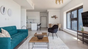 Una sala moderna con un sofá color turquesa, una mesa de centro de madera y una silla negra. En el fondo, se ve una cocina con armarios blancos y un comedor.