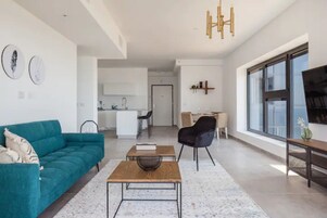 Uma sala de estar moderna com um sofá verde-água, uma mesa de centro de madeira e uma cadeira preta. Dá para ver uma cozinha ao fundo, com armários brancos e uma área de jantar.