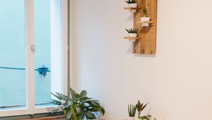 Une pièce chaleureuse avec une table blanche, deux chaises, une petite table d’appoint en bois sur laquelle se trouve une bougie, et un canapé blanc. Trois plantes en pot sont disposées sur une étagère en bois au-dessus de la table.