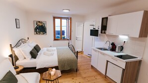 Une chambre compacte comprenant un lit, une table de chevet et une fenêtre. À côté se trouve une petite cuisine équipée d’un évier, d’une cuisinière et de placards.