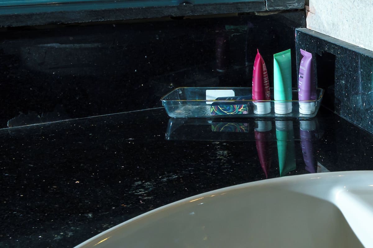 Una encimera de baño con superficie de mármol negro, una bandeja de vidrio transparente con tres tubos de cosméticos y un lavamanos blanco.