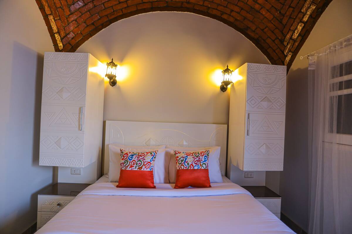 Una cama con sábanas blancas y dos almohadas rojas, cabecera blanca y dos apliques de pared.