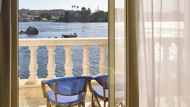 A balcony with a view of a river, a boat, and distant buildings. - NUB INN (Aswan)