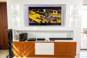 A modern living room with a flat-screen TV mounted on a white wall, a wooden entertainment unit below, and a black speaker to the left.