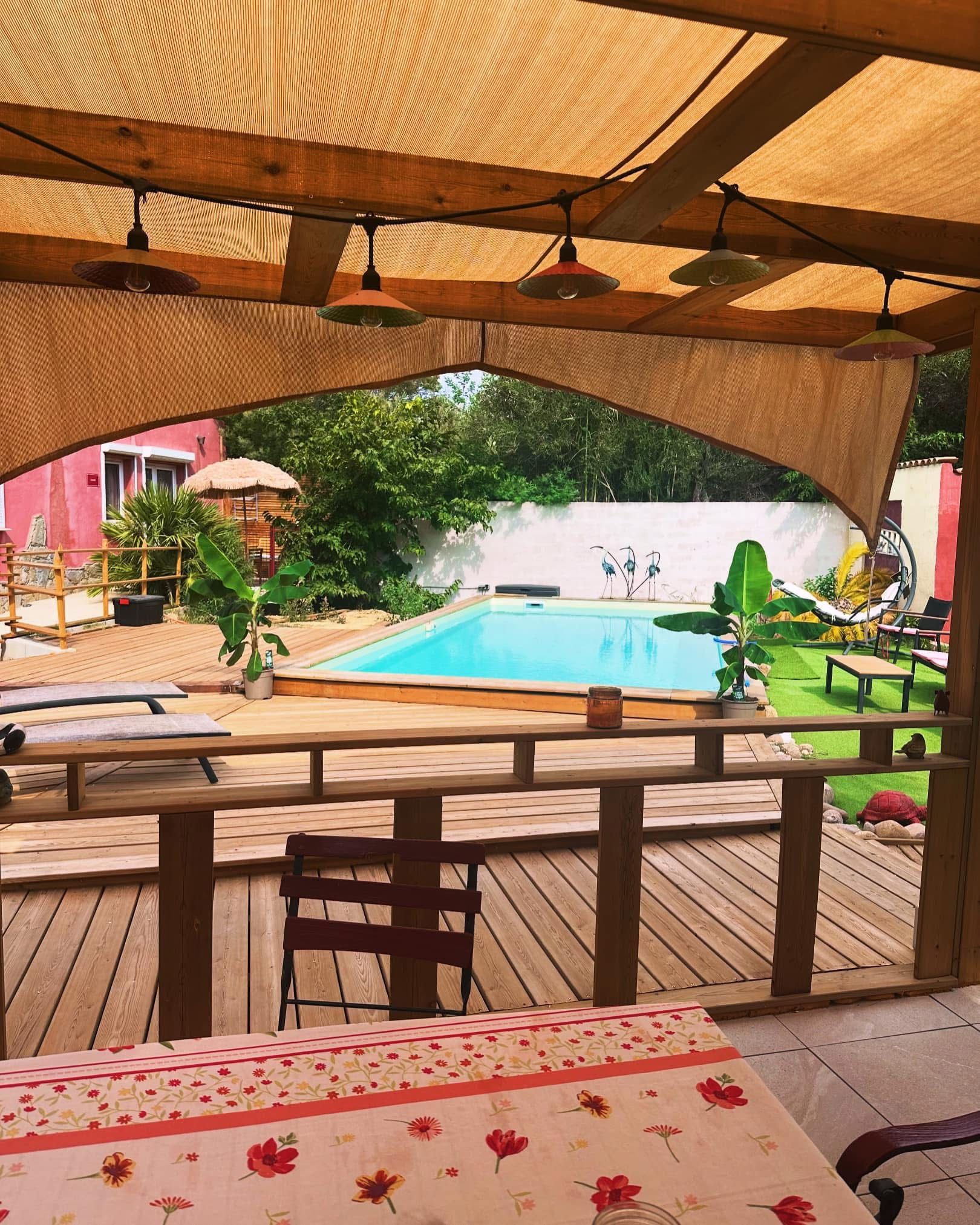 Une terrasse en bois avec une piscine, entourée de verdure et dotée d’un espace couvert avec des tables et des chaises.