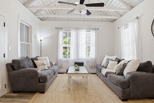 A room with two grey sofas, a white coffee table, a ceiling fan, and large windows with white curtains.