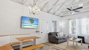 Une pièce avec un plafond blanc, un ventilateur de plafond, un lustre, un canapé, une table basse, un banc, un tapis à motifs et un téléviseur diffusant une scène de plage.