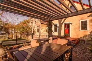 Um pátio externo com mesa e cadeiras de madeira, uma pérgula e um edifício com porta vermelha.