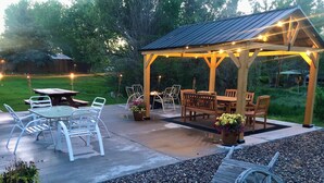Un patio al aire libre con un porche de madera, sillas de metal blanco, una mesa de picnic y una rueda de madera decorativa.