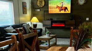 Una sala de estar con una mesa de comedor de madera, un televisor mostrando una escena de equitación, un sofá, una lámpara y un reloj de pared.