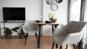 Una sala de estar moderna con un televisor de pantalla plana, dos sillones grises, una mesita redonda con una planta y una ventana con persianas.