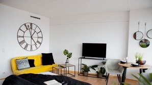 Una sala moderna con un sofá amarillo, un reloj de pared grande, un televisor de pantalla plana y una zona de comedor con una mesa pequeña y sillas.