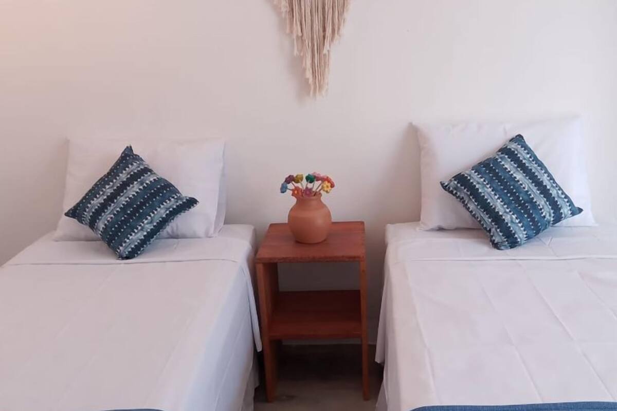 A room with two beds, each with a white bedspread and a blue blanket. There is a wooden nightstand with a vase of flowers between the beds.