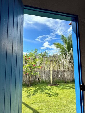 A view through a blue-framed door into a grassy yard with a bamboo fence, a palm tree, and a clear sky. - Le Petit Suítes Praia de Lages (Porto de Pedras)