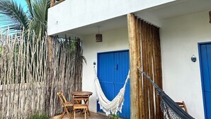 Un bâtiment à deux étages, avec une façade blanche, une porte bleue et un balcon en bois. On y trouve un hamac, une chaise en bois et une petite table sur la terrasse.