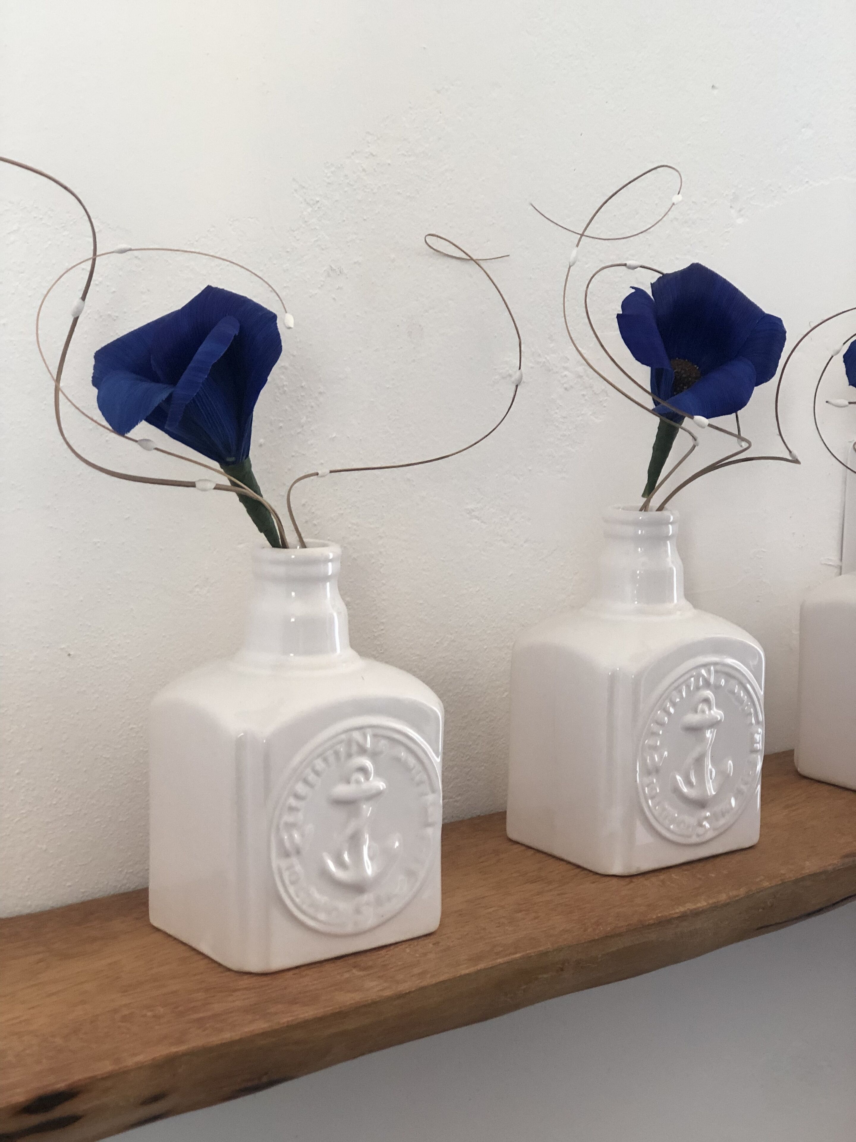 Tre vasi di ceramica bianca con decorazioni a rilievo, ognuno con un singolo fiore blu, disposti su una mensola di legno davanti a un muro spoglio.