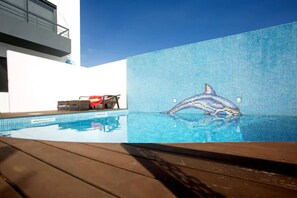 Ein Swimmingpool mit einem Delfin-Mosaik, einer Holzterrasse und einem Liegestuhl mit einem roten Handtuch.