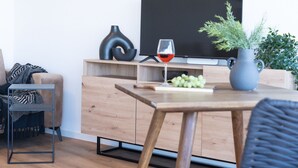 Una sala de estar moderna con piso de madera, un televisor sobre un mueble de madera, una mesa de comedor con un florero y uvas, y una silla.