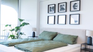 Un dormitorio con una cama grande, ropa de cama verde, una planta en maceta, una mesita de noche y una pared con cuadros enmarcados.