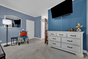 Ein Schlafzimmer mit einem Bett, einem Schminktisch, einem Sessel, einer Lampe und einem an der Wand befestigten Fernseher.