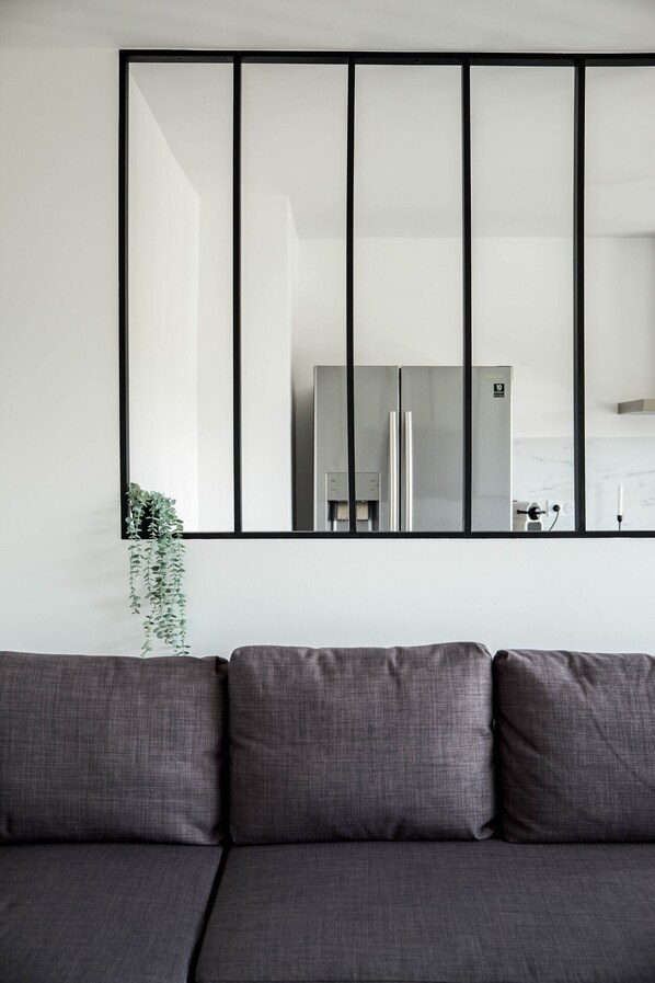 A modern living room with a large mirror reflecting a kitchen area, a grey sofa, and a hanging plant.