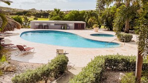 Una piscina con borde curvo, rodeada por una zona de patio con sillones de descanso y un pequeño edificio con techo plano.