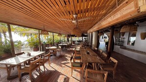Un área de comedor al aire libre con mesas y sillas de madera, plantas en macetas y techo de madera.