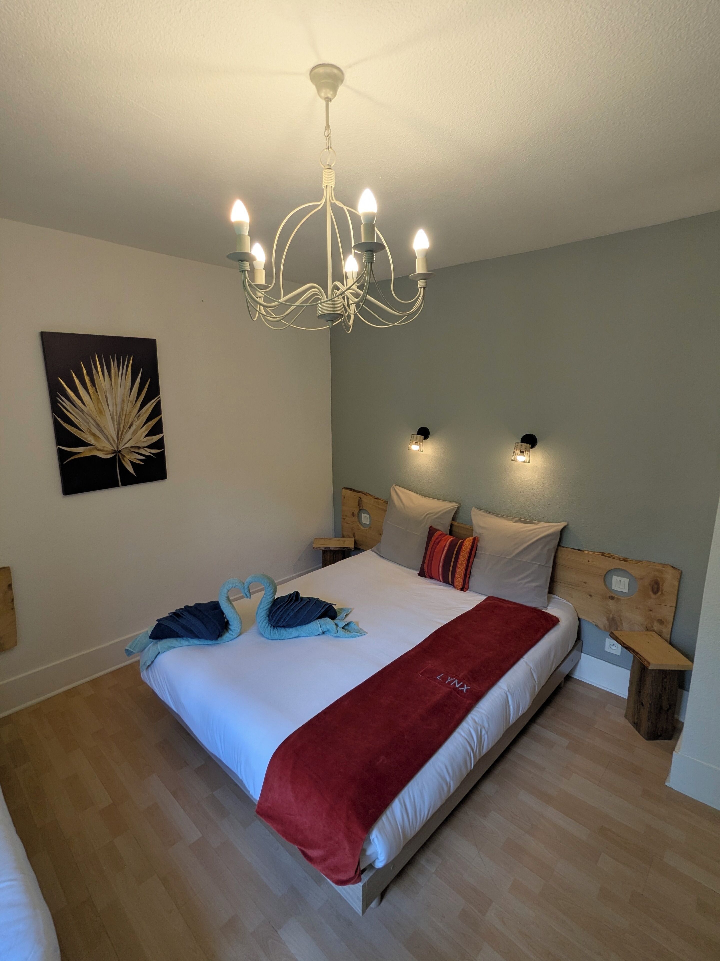 A bedroom with a bed, a chandelier, wall-mounted lights, a wooden headboard, and a wall art piece.