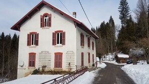 Une maison à deux étages avec des volets rouges et un toit enneigé, entourée d’arbres et d’un paysage hivernal.