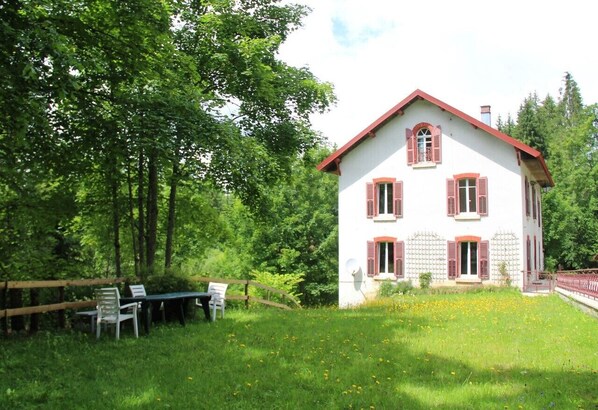 Une maison blanche aux volets rouges, entourée d’une clôture en bois et d’un espace verdoyant avec une table et des chaises.